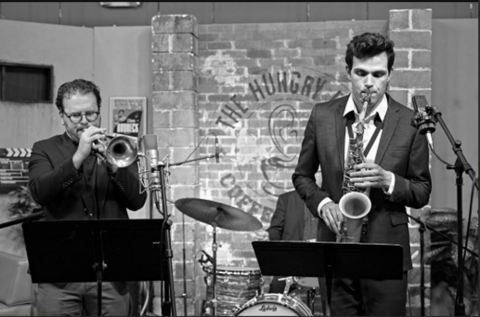 A black and white photograph of 2 musicians, 1 playing a trumpet and 1 playing a saxophone.